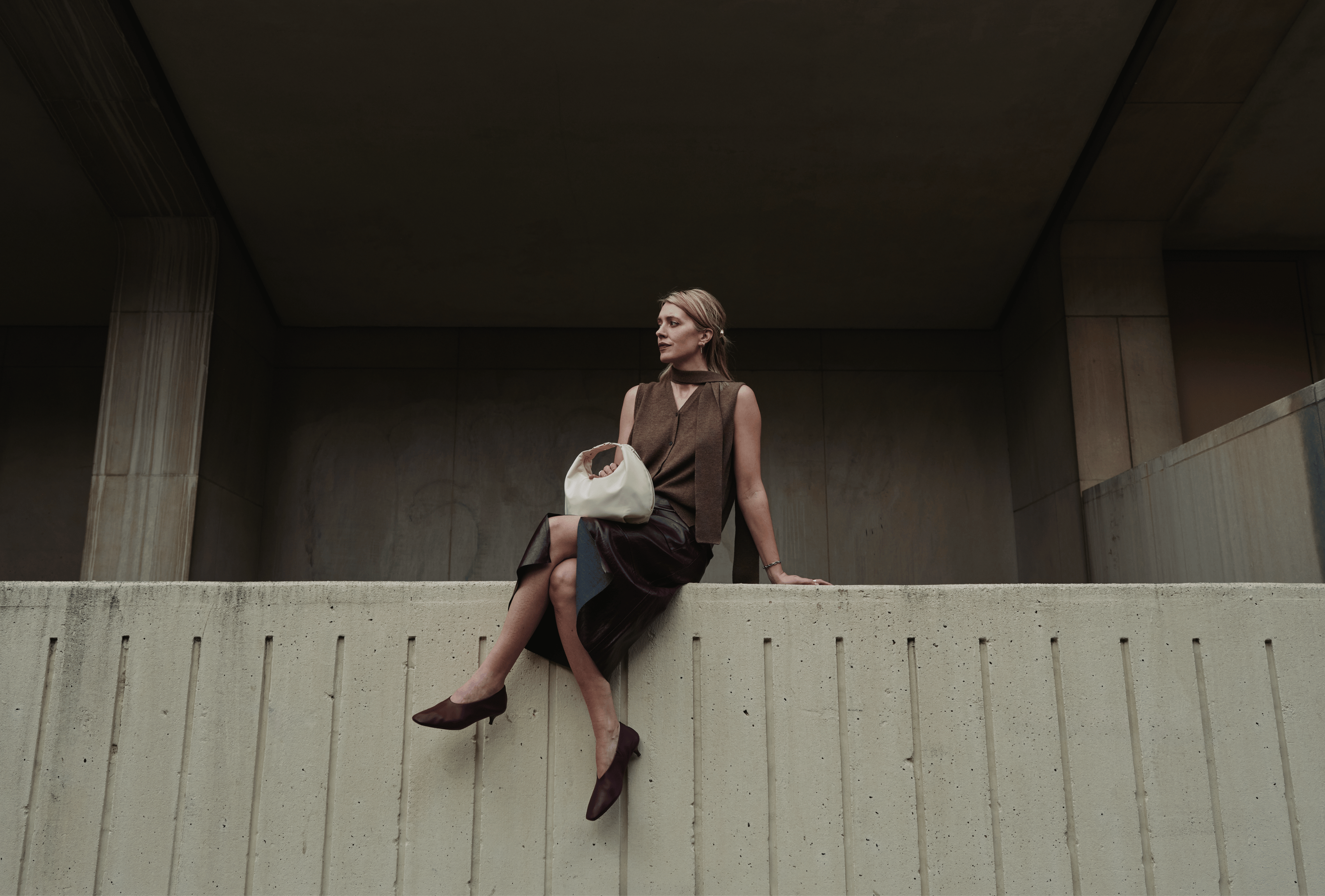 Woman sitting on a ledge in urban setting with a Lo & Sons Aoyama crossbody handbag, wearing a brown dress and shoes.