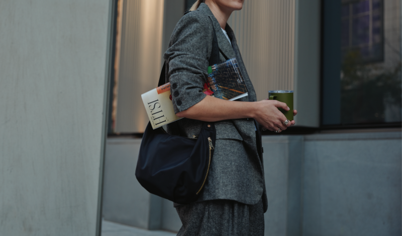 Woman holding a black Lo & Sons Aoyama nylon crossbody bag and a green drink, walking past a building in an urban setting.