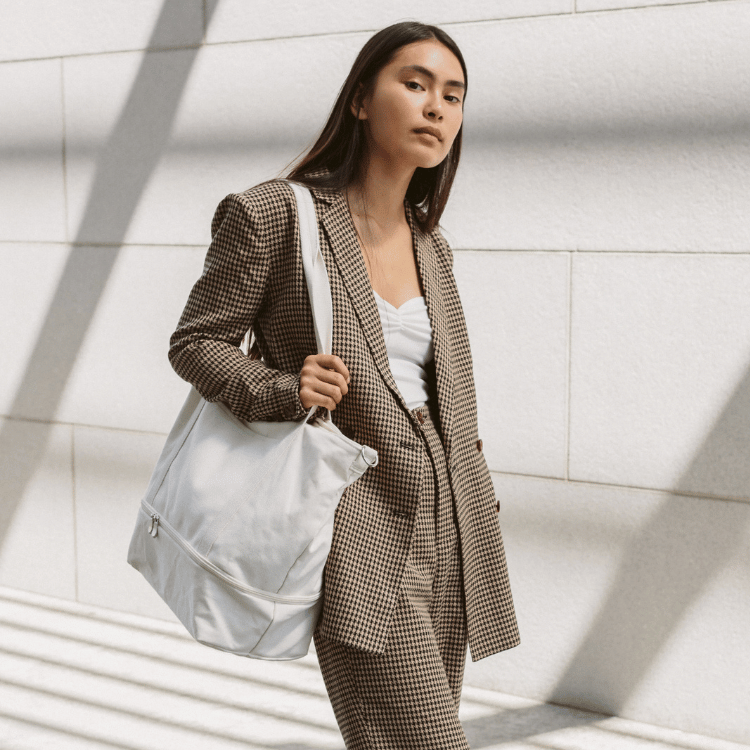 Woman in a checkered suit blazer with pants holding a natural colored Lo & Sons Catalina Deluxe Tote bag against a light-colored wall.