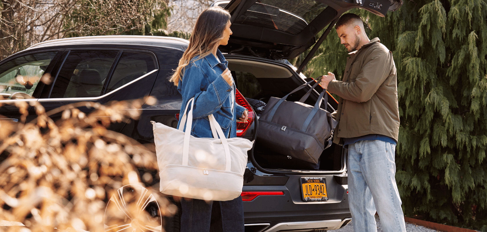 Two people with Lo & Sons Catalina Deluxe weekender bags near an open car packing the trunk for a trip