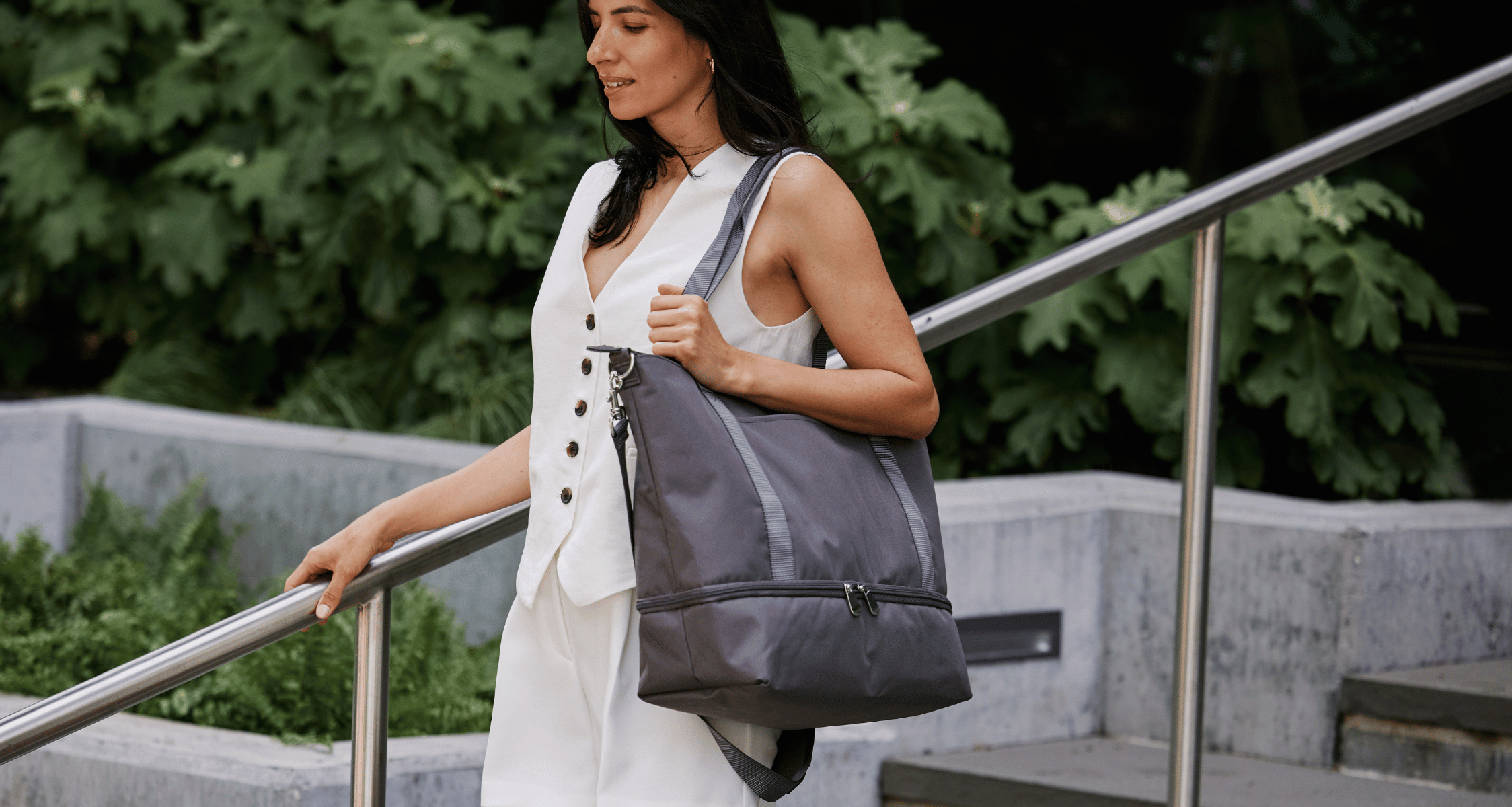 Woman holding a gray Lo & Sons Catalina Deluxe Tote bag outside an office building for work with greenery in the background