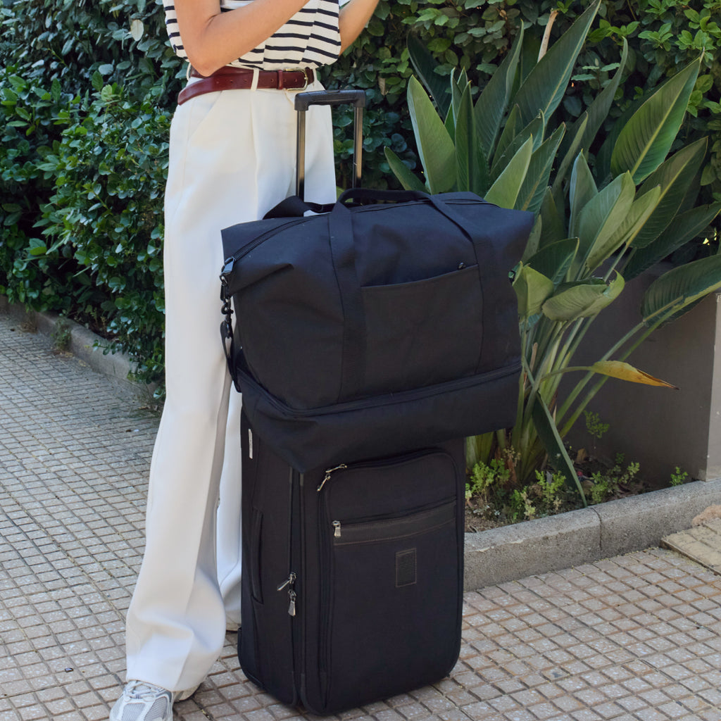 Woman standing outdoors holding a Lo & Sons Catalina Deluxe in black poly that's sitting on top of a black suitcase.