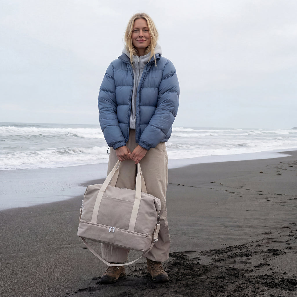woman in puffer jacket and hoodie on a beach in the winter time holding the Lo & Sons Catalina Deluxe in Dove Grey canvas 