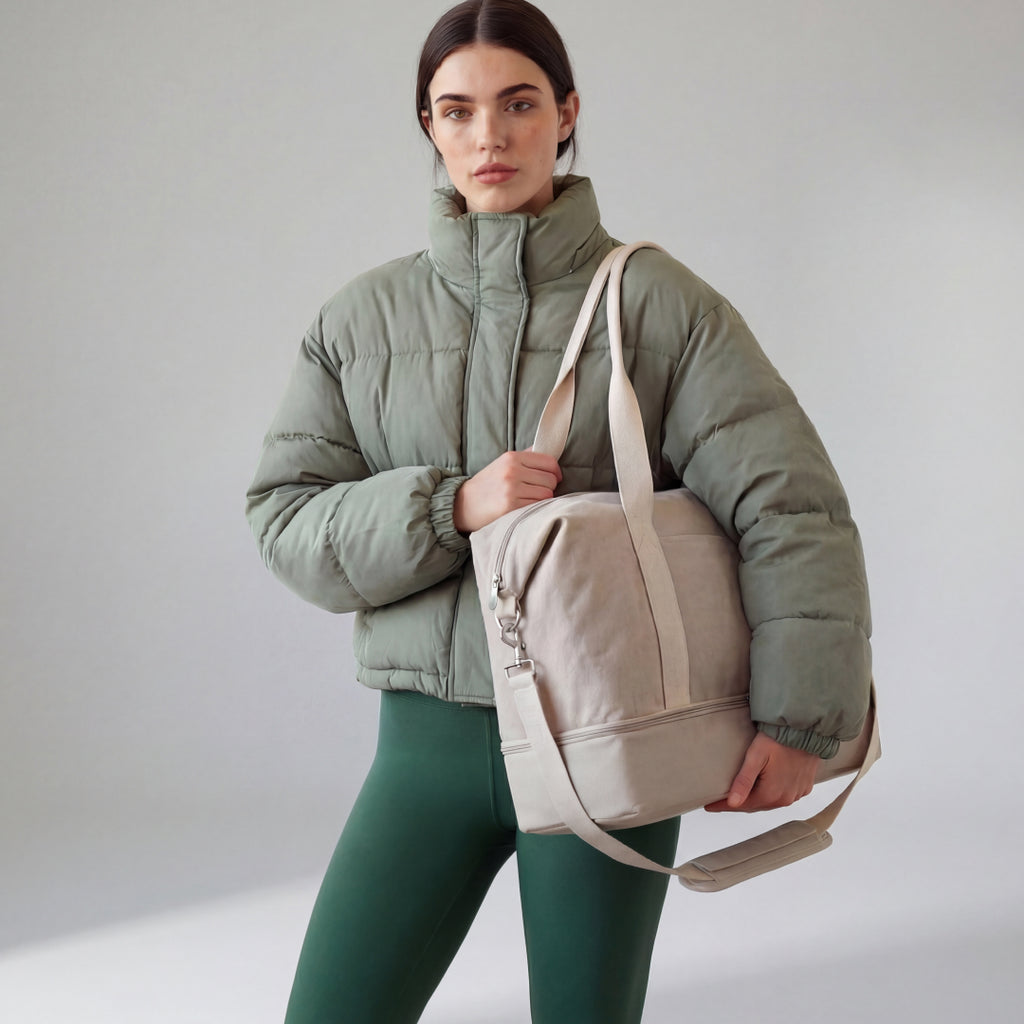 Woman carrying a Lo & Sons Catalina Deluxe in Dove Grey canvas, wearing a green puffer jacket and leggings against a plain background