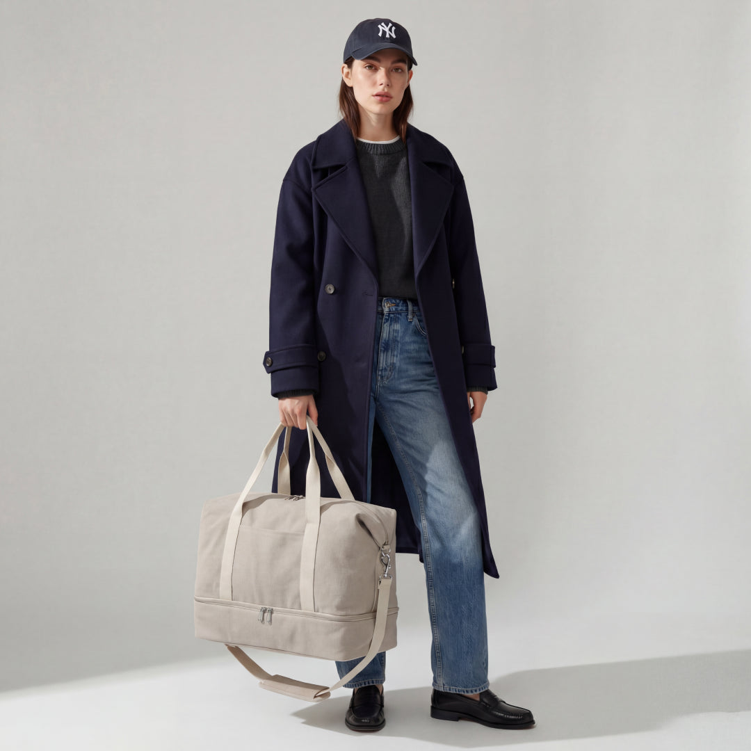 Woman carrying the Lo & Sons Catalina Deluxe in Dove Grey Canvas, wearing a navy coat and blue jeans