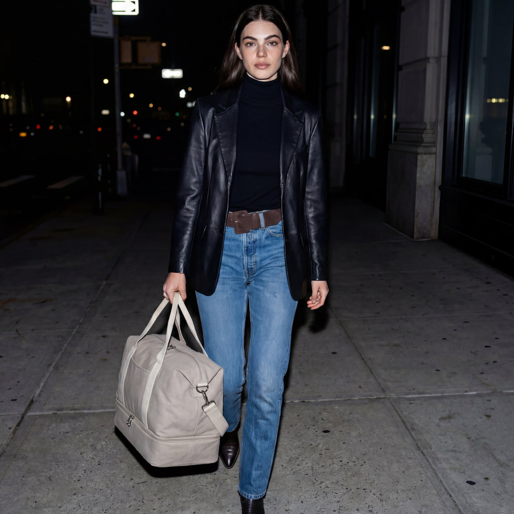 Woman walking on a city street at night, holding a Lo & Sons Catalina Deluxe dove grey duffel bag.