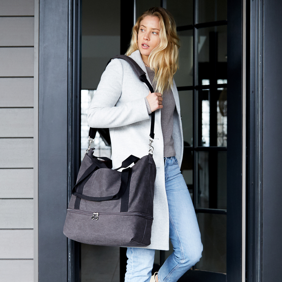 Woman holding a Lo & Sons Catalina Deluxe Tote in color midnight ash as she exits a home about to go on a trip