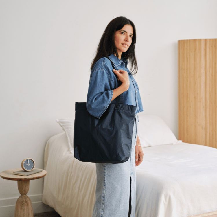 Woman holding a navy blue Lo & Sons Del Mar packable reusable tote bag in a bedroom setting getting ready to run errands for the day