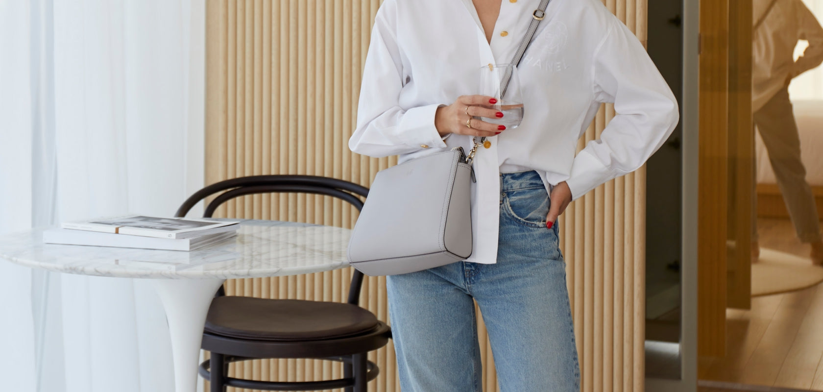 woman in bedroom carrying the Lo & Sons Pearl crossbody bag in light grey saffiano