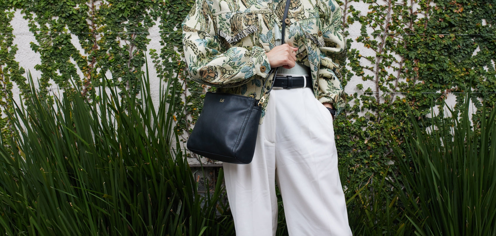 woman standing in front a wall with plants carrying the Lo & Sons Pearl crossbody bag in black nappa leather