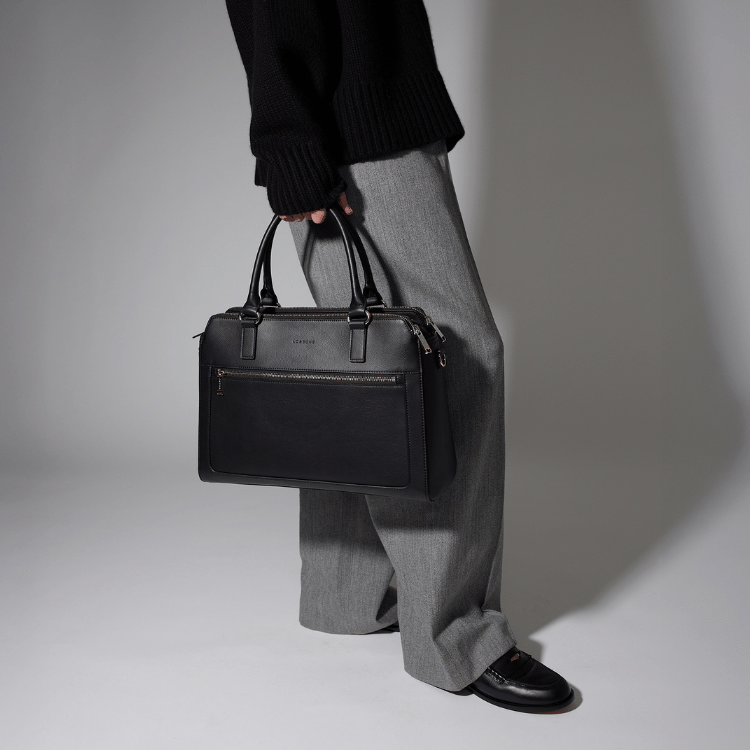 Woman holding a Lo & Sons black leather Trinity laptop tote bag against a neutral background