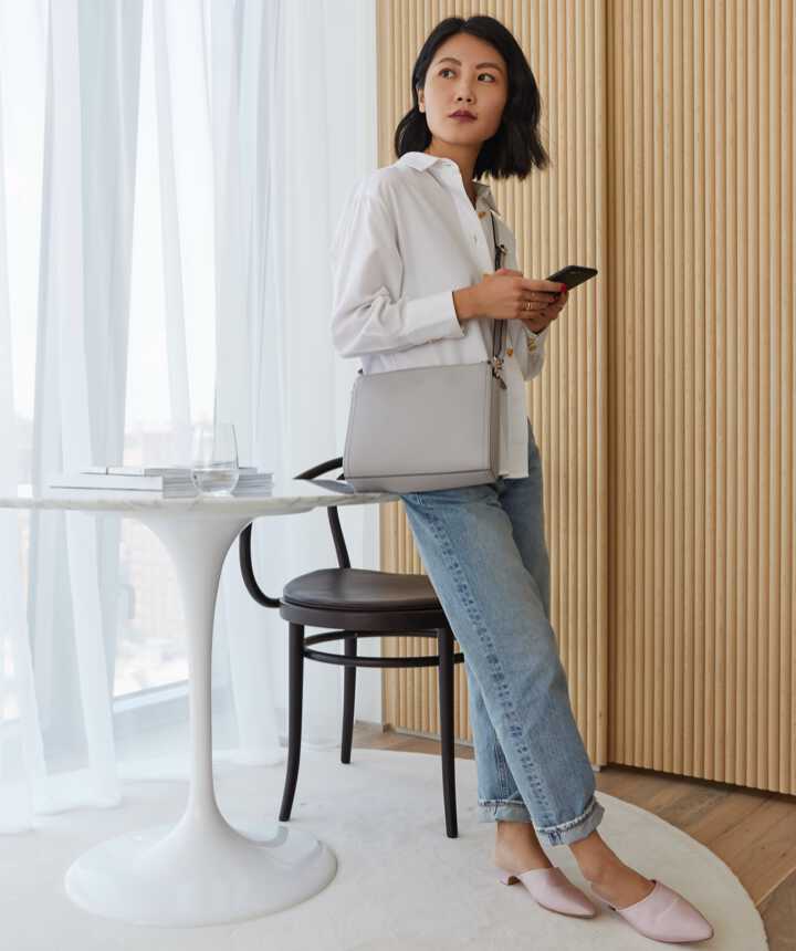 woman in hotel room carrying the Lo & Sons Pearl bag in light grey saffiano as a crossbody bag